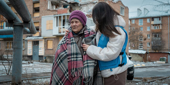 Olena, 85, stands outside her damaged home in Kharkiv, Ukraine, supported by humanitarian aid after a May 2024 attack.