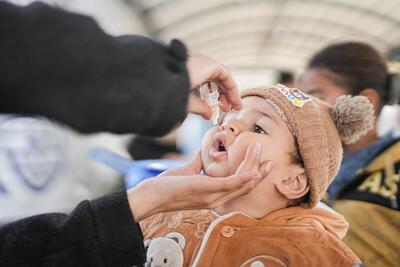 A healthcare worker administers an oral polio vaccine to a young child wearing a brown coat and a knitted hat with pom-poms. Another caregiver is visible in the background.
